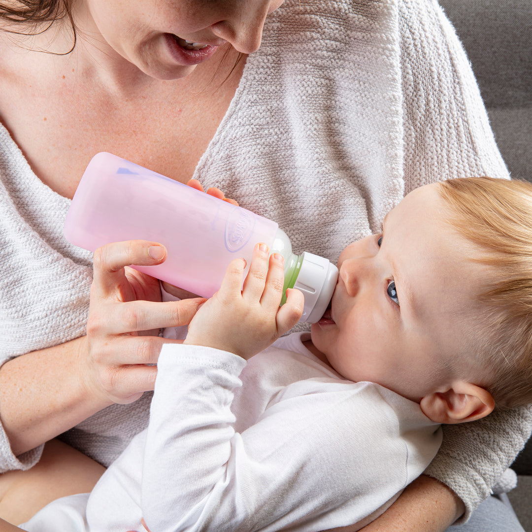 A baby contentedly drinks from a pink Dr. Brown’s Natural Flow® Options+™ Narrow Glass Bottle Silicone Sleeve, held by an adult wearing a light sweater. The little one, dressed in white, appears perfectly at ease indoors, possibly nestled on a cozy couch.