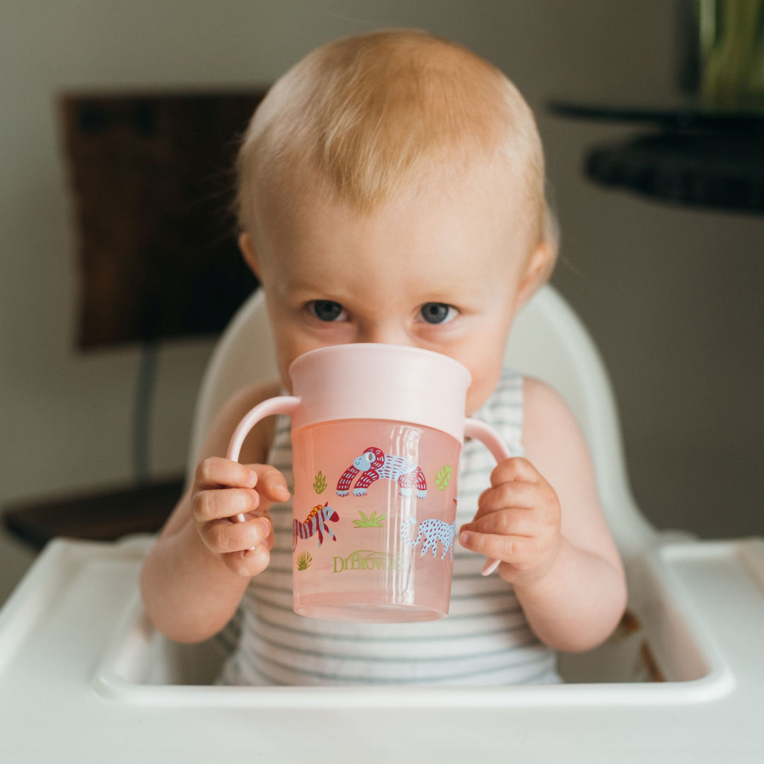 A baby is seated in a high chair, sipping happily from a blue Cheers360™ cup by Dr. Brown's, designed for mess-free learning. Wearing a striped sleeveless shirt, the little one glances toward the camera while enjoying their leak-free drinking experience.