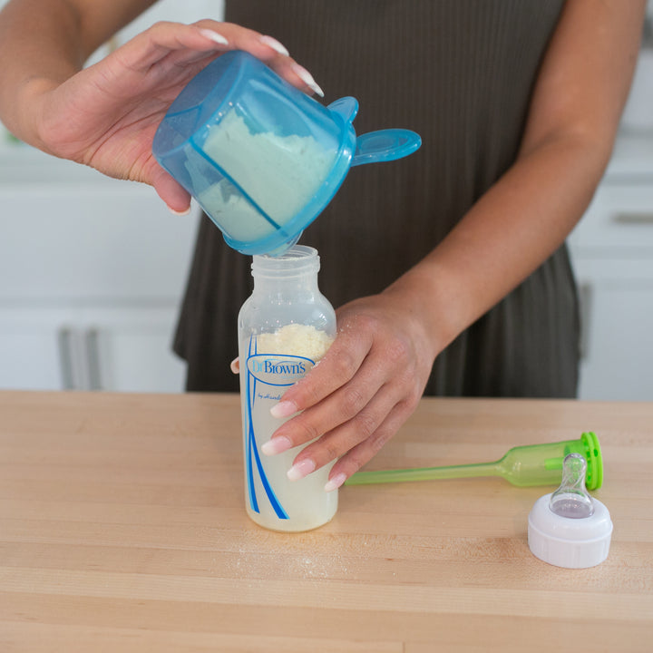 A person uses a blue Dr. Brown's® Baby Formula Dispenser to pour powdered formula into a baby bottle on a wooden countertop. Nearby, a green funnel and the lid accompany the 9 oz bottle, all set for efficient baby feeding.