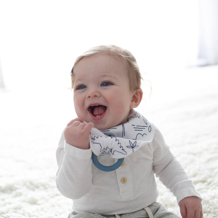 A smiling baby sits on a soft, white surface, wearing Dr. Brown’s™ Bandana Bib with a blue design and a long-sleeved white shirt. The teething baby, with short light brown hair and blue eyes, looks upwards with one hand raised to their mouth while bright lighting fills the room.