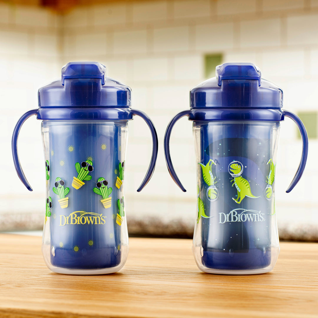 Two Dr. Brown’s Milestones™ Insulated Straw Cups with blue lids and handles rest on a wooden surface. One spill-proof cup showcases a cactus design, while the other features a dinosaur pattern. The background displays a tiled wall and a blurred kitchen setting.