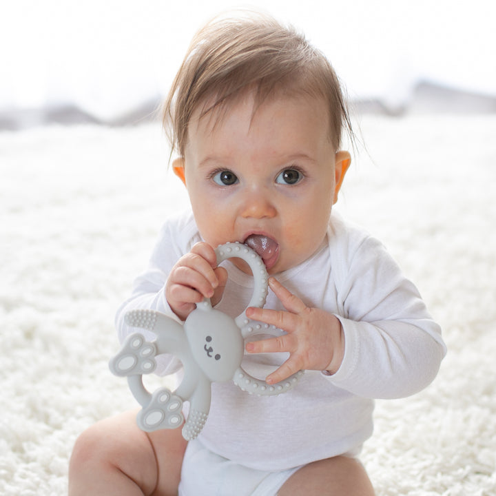 A baby dressed in a white outfit is seated on a soft, light-toned carpet, holding and chewing on Dr. Brown's™ Flexees™ Bunny Teether. The silicone teether provides soothing relief for teething, while the softly blurred background enhances the cozy and warm ambiance.