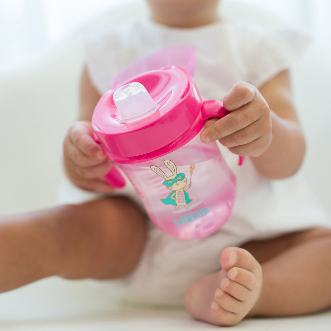 A baby, dressed in a white outfit, sits on a white surface holding Dr. Brown’s® Soft-Spout Toddler Cup in pink featuring a cartoon bunny design. Ideal for transitioning from bottle to cup, the cup's silicone spout ensures spill-free learning.