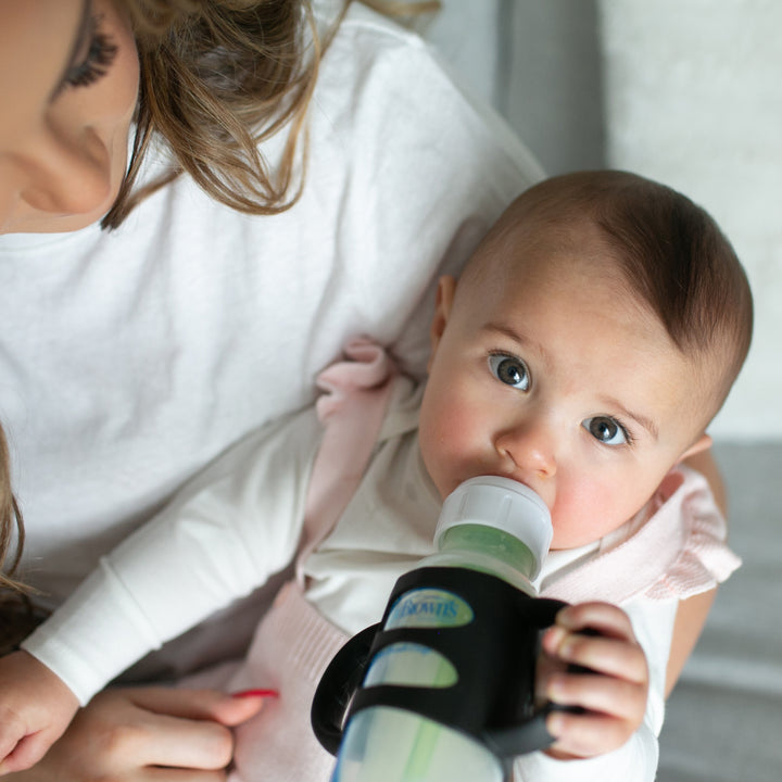 A blue-eyed baby is comfortably nestled in an adult's arms, confidently demonstrating its independent drinking skills with a bottle featuring Dr. Brown’s® Milestones™ Narrow Silicone Handles. The baby, clad in a long-sleeved shirt, gazes directly at the camera while the partially visible adult wears a white top.