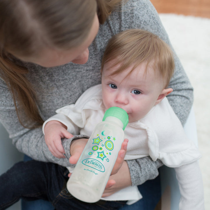 A woman with long hair is comfortably feeding a baby dressed in a white shirt using Dr. Brown's Natural Flow® Anti-Colic Options+™ Narrow Sippy Bottle Starter Kit, 8oz/250mL. The bottle, adorned with colorful designs and featuring a green top, ensures an anti-colic experience as she gently holds the baby while wearing a cozy gray sweater.