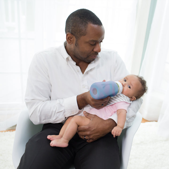 A man in a white shirt sits in a chair, gently holding a baby. He is feeding the baby with a glass bottle featuring Dr. Brown's Natural Flow® Options+™ Wide-Neck Glass Bottle Silicone Sleeves. The room is softly lit, creating a peaceful atmosphere.