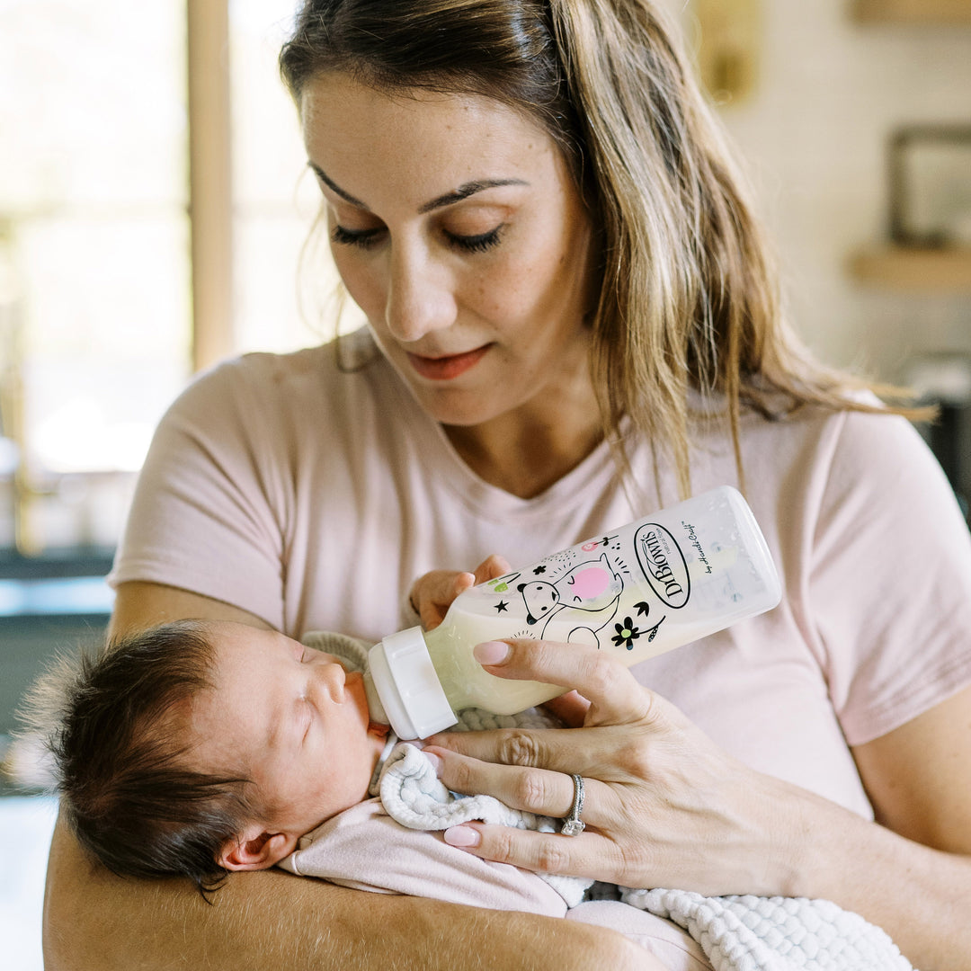 A woman with long hair, dressed in a pink shirt, is gently feeding her baby using Dr. Brown’s Natural Flow® Anti-Colic Options+™ Narrow Baby Bottle 8oz/250mL. The baby, nestled in a blanket, relaxes peacefully in her arm within the cozy home environment softly illuminated.