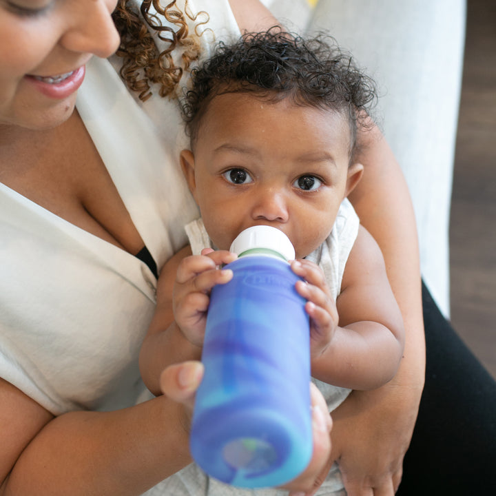 A baby with curly hair is drinking from a blue Dr. Brown’s Natural Flow® Options+™ Narrow Glass Bottle, which has a silicone sleeve for protection. The baby is being held by an adult, who is partially visible, as they sit in a cozy environment. The baby is dressed in a light-colored outfit.