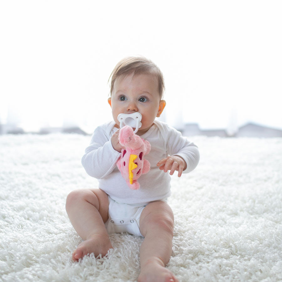 A baby wearing a white onesie is seated on a fluffy white carpet, holding a pink stuffed toy designed as Dr. Brown’s® Lovey Pacifier and Teether Holder, Dinosaur. The soothing ambiance is completed by the Dr. Brown's HappyPaci resting comfortably in its mouth, set against a softly lit background that enhances the serene atmosphere.