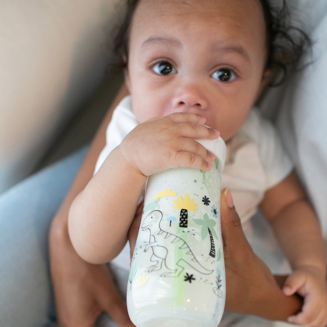 A baby in a white shirt, held gently, drinks from a Dr. Brown’s Natural Flow® Anti-Colic Options+™ Wide-Neck Baby Bottle, featuring dinosaur illustrations. The wide-neck design ensures comfort as the baby gazes upward with wide eyes.