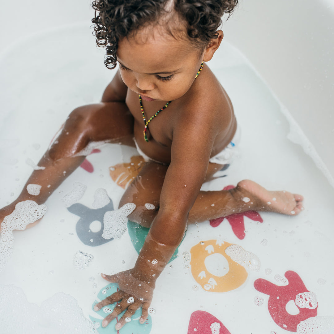 A toddler sits in a bathtub filled with water and suds, safely playing with colorful foam letters that float around. The child, with curly hair and a beaded necklace, enjoys bath time on Dr. Brown’s™ CleanUp™ Sit & Splash Non-Slip Bath Stickers in the plain white tub for extra safety.