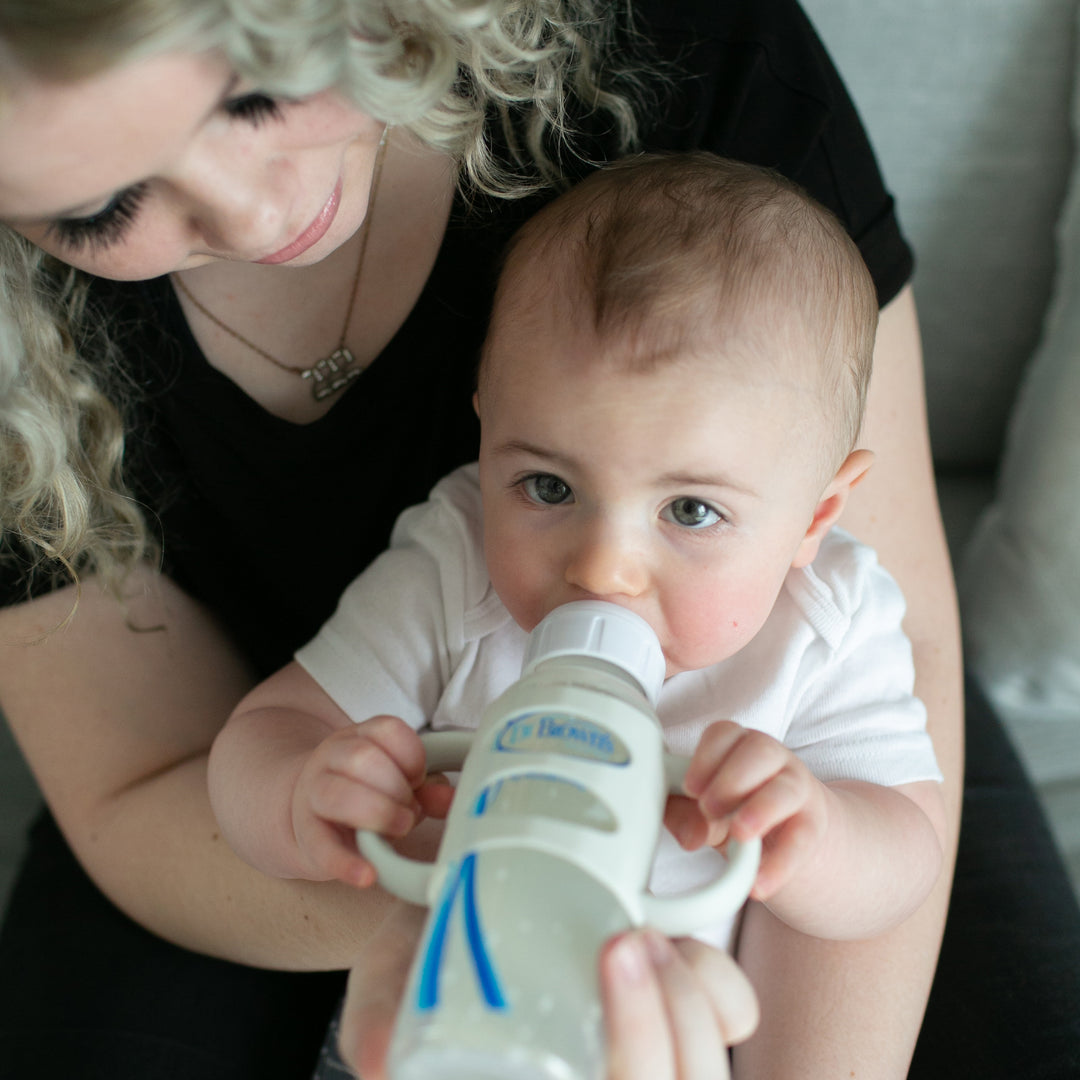 A woman with curly hair holds a baby who is drinking from a Dr. Brown’s Narrow Bottle equipped with Dr. Brown’s® Milestones™ Narrow Silicone Handles. The baby, dressed in a white shirt and practicing independent drinking skills, looks up while sipping, encapsulating a nurturing moment.