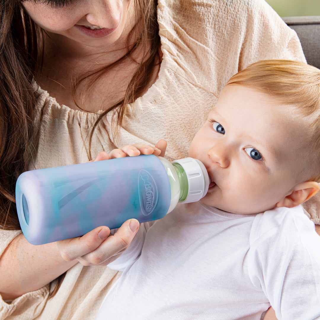 A baby in a white shirt is drinking from a Dr. Brown's Natural Flow® Options+™ Narrow Glass Bottle, which features a blue and purple silicone sleeve, held by someone with long brown hair wearing a light beige top. They lounge on the couch as the protective sleeve ensures safe and comfortable feeding.