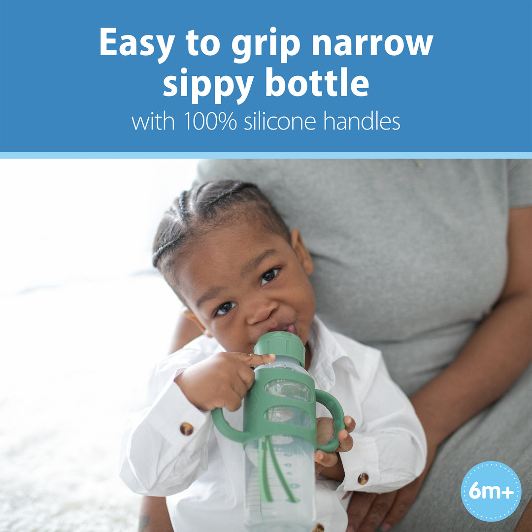 A toddler in a white shirt drinks from a Dr. Brown’s® Milestones™ Narrow Sippy Spout Bottle with Silicone Handles, sitting on an adult's lap. Text above reads, "Easy to grip narrow sippy bottle with 100% silicone handles." A "6m+" label is in the corner.