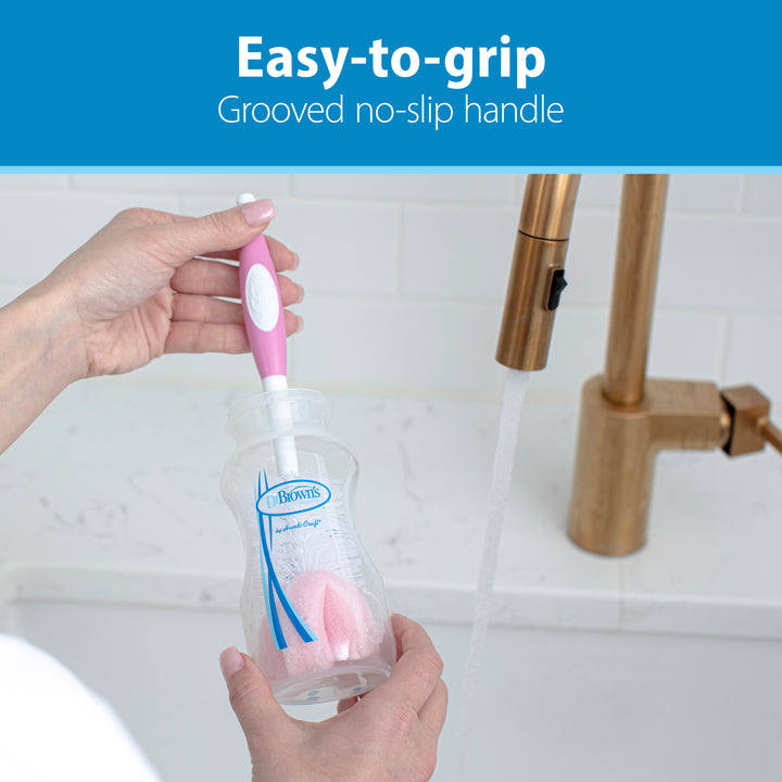 A beloved parent moment: washing a baby bottle with Dr. Brown’s Natural Flow® Baby Bottle Brush, featuring its distinct pink and white design, under a running kitchen faucet. The easy-to-grip grooved no-slip handle from Dr. Brown's makes the task effortless, and the built-in nipple cleaner guarantees thorough cleanliness every time.