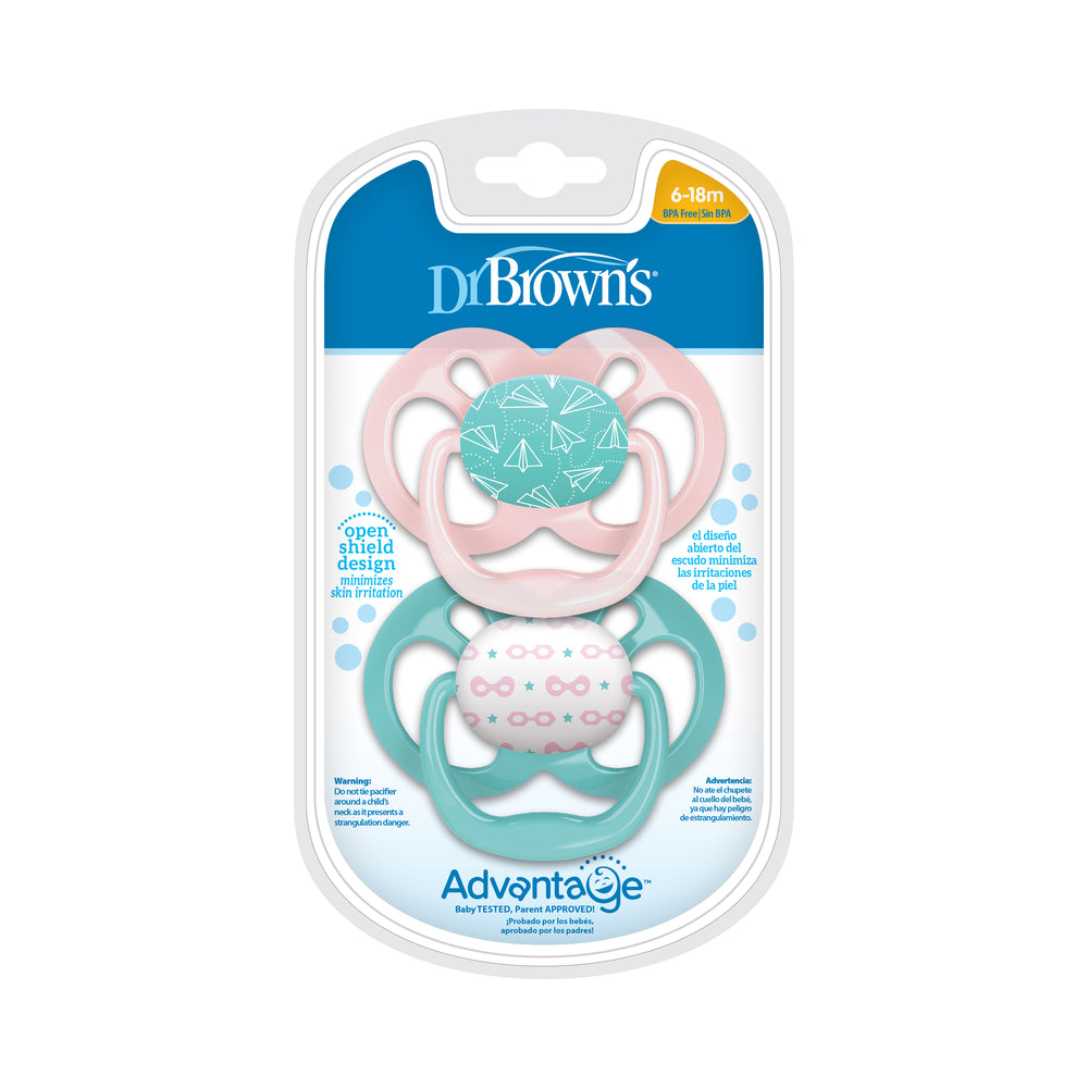 Product packaging for Dr. Brown’s™ Advantage™ Pacifiers, 2 count, includes two open shield design soothers. The top pink pacifier features a paper airplane pattern, while the bottom teal one showcases a heart and triangle design. These pacifiers have a symmetrical, reversible bulb and are suitable for ages 6-18 months.