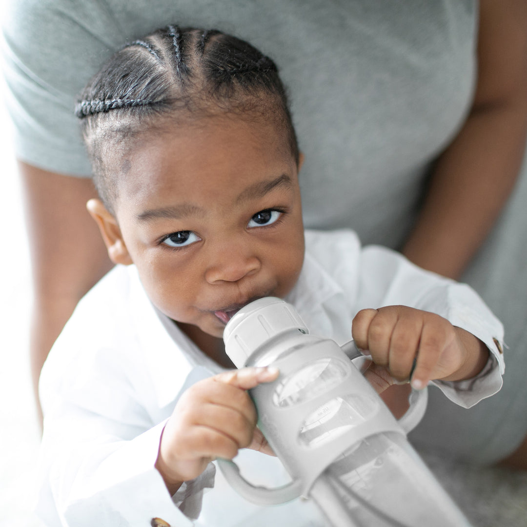 A toddler with braided hair is drinking from a gray Dr. Brown's® Milestones™ Narrow Sippy Spout Bottle featuring silicone handles, 8 oz/250 mL. The child, dressed in a white shirt, is being held by an adult wearing a gray shirt and gazes up at the camera with bright eyes.