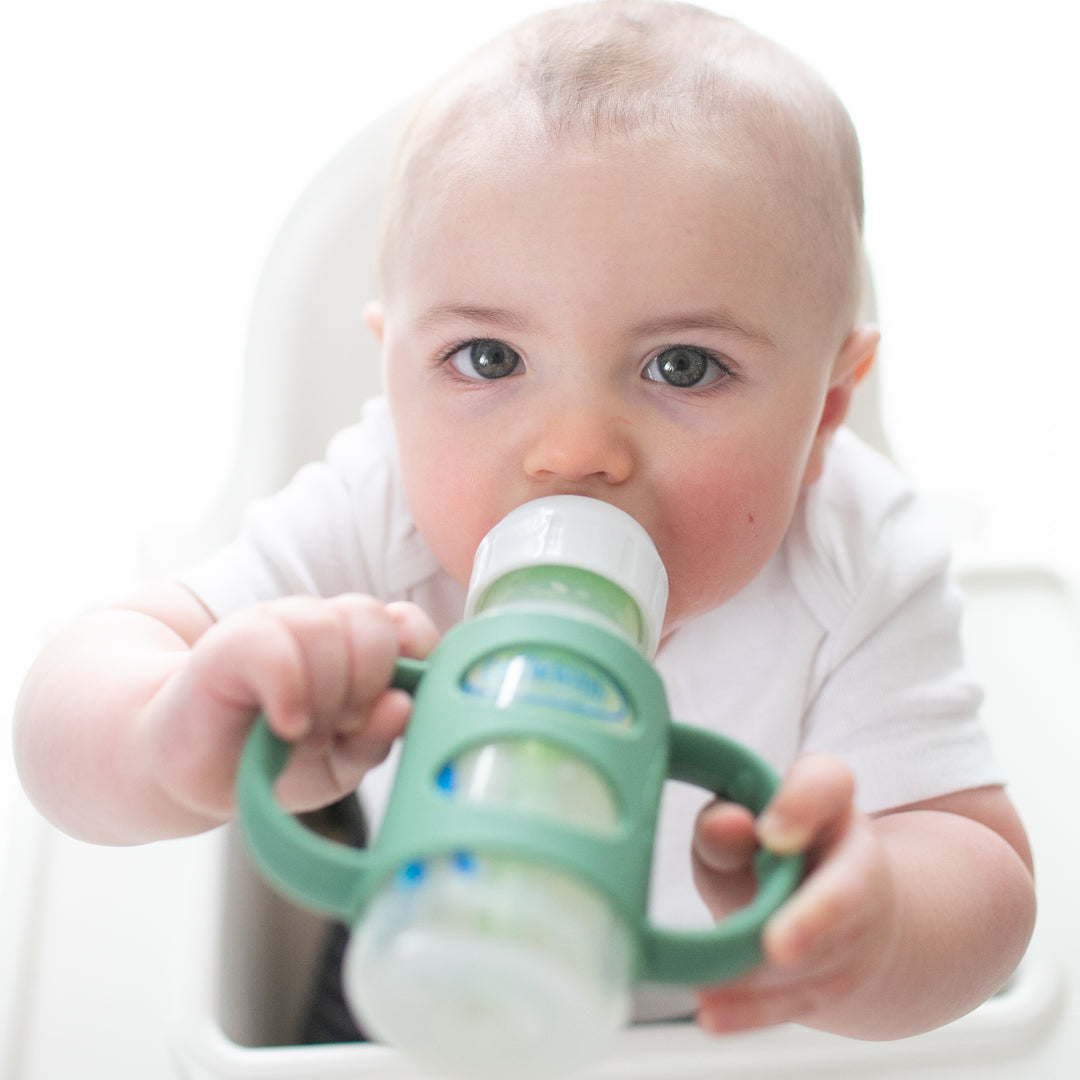 A baby with short hair is seated in a high chair, holding Dr. Brown’s® Milestones™ Narrow Silicone Handles, ideal for transitioning from bottle to cup. Dressed in a white shirt and gazing directly at the camera, the baby demonstrates emerging independent drinking skills against a softly blurred, mostly white background.