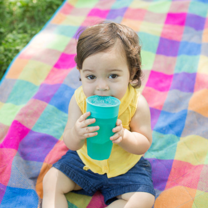 A child in a yellow shirt sits on a colorful checkered blanket outdoors, holding a Dr. Brown's® Milestones™ Cheers360™ Cup, featuring a 360-degree design with a Sip and See silicone valve. The lush green grass surrounding the blanket creates a cozy and vibrant setting for the little one's refreshing sip.