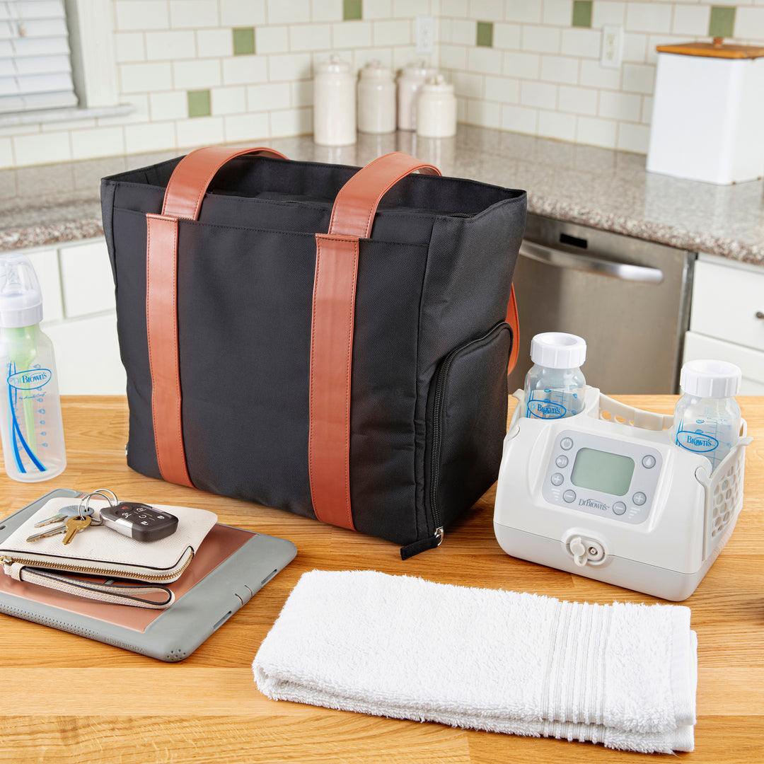 A sleek black Dr. Brown’s Breast Pump Carryall Tote Bag with brown handles is placed on the kitchen countertop, surrounded by essentials like a breast pump, baby bottles, a tablet, and keys. Ideal for mom time, its stylish design blends seamlessly against the backdrop of kitchen tiles and appliances.