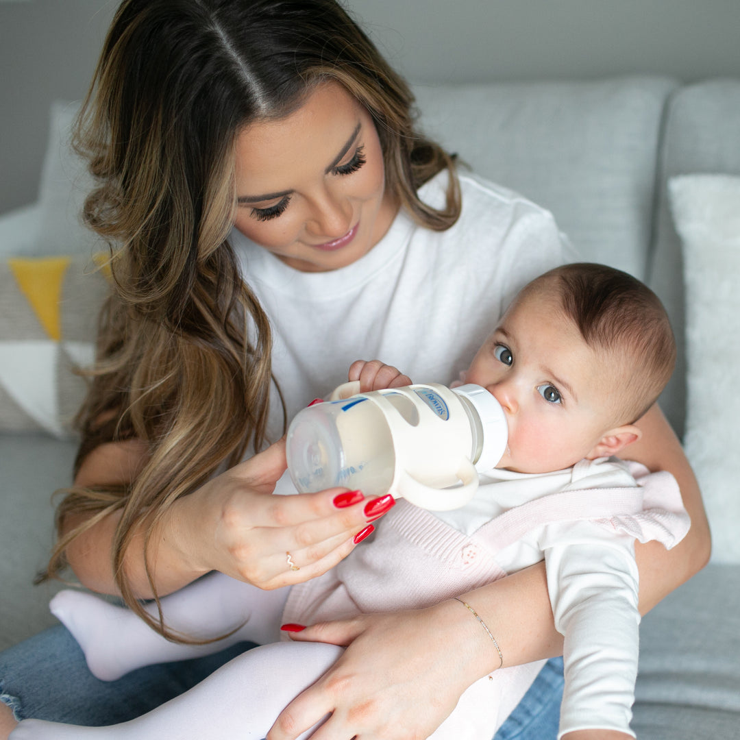 A woman with long hair, wearing a white shirt, is sitting on a couch in a cozy living room, bottle-feeding a baby in her arms. The baby contentedly gazes up while drinking from Dr. Brown’s® Milestones™ Wide-Neck Silicone Handle bottles by Dr. Brown's.