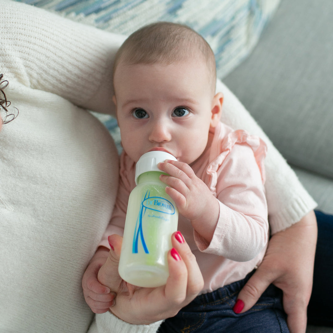 A baby in a pink shirt is being held by an adult, drinking from a Dr. Brown’s Natural Flow® Anti-Colic Options+™ Narrow Glass Baby Bottle, which features a Level 1 Slow Flow Nipple designed to mimic the breast. The adult's hand supports the bottle containing milk, while the baby has a focused expression. The adult is dressed in a white sweater.