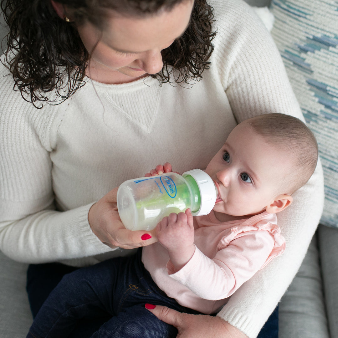 A woman in a white sweater enjoys a peaceful feeding experience on the couch, sitting relaxed with a baby in her lap. The baby, wearing a pink top, looks up contentedly while drinking from a bottle featuring Dr. Brown’s Natural Flow® Wide-Neck Baby Bottle Silicone Nipple by Dr. Brown's.