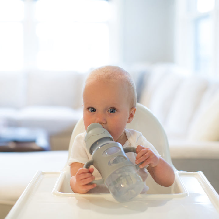 A baby sits in a high chair, confidently holding the Dr. Brown’s™ Milestones™ Wide-Neck Sippy Straw Bottle with Silicone Handles, 9 oz/270 mL, drinking from it with both hands. The background reveals a softly blurred living room with a cream-colored sofa, reflecting the little one's growing independent drinking skills.