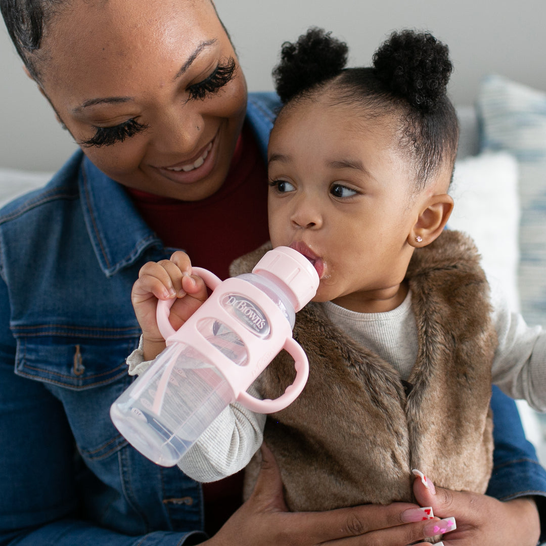 A woman, with neatly styled hair and donned in a denim jacket, smiles at a toddler wearing a fur vest. The toddler is drinking from Dr. Brown's® Milestones™ Narrow Sippy Spout Bottle with Silicone Handles, 8 oz/250 mL.