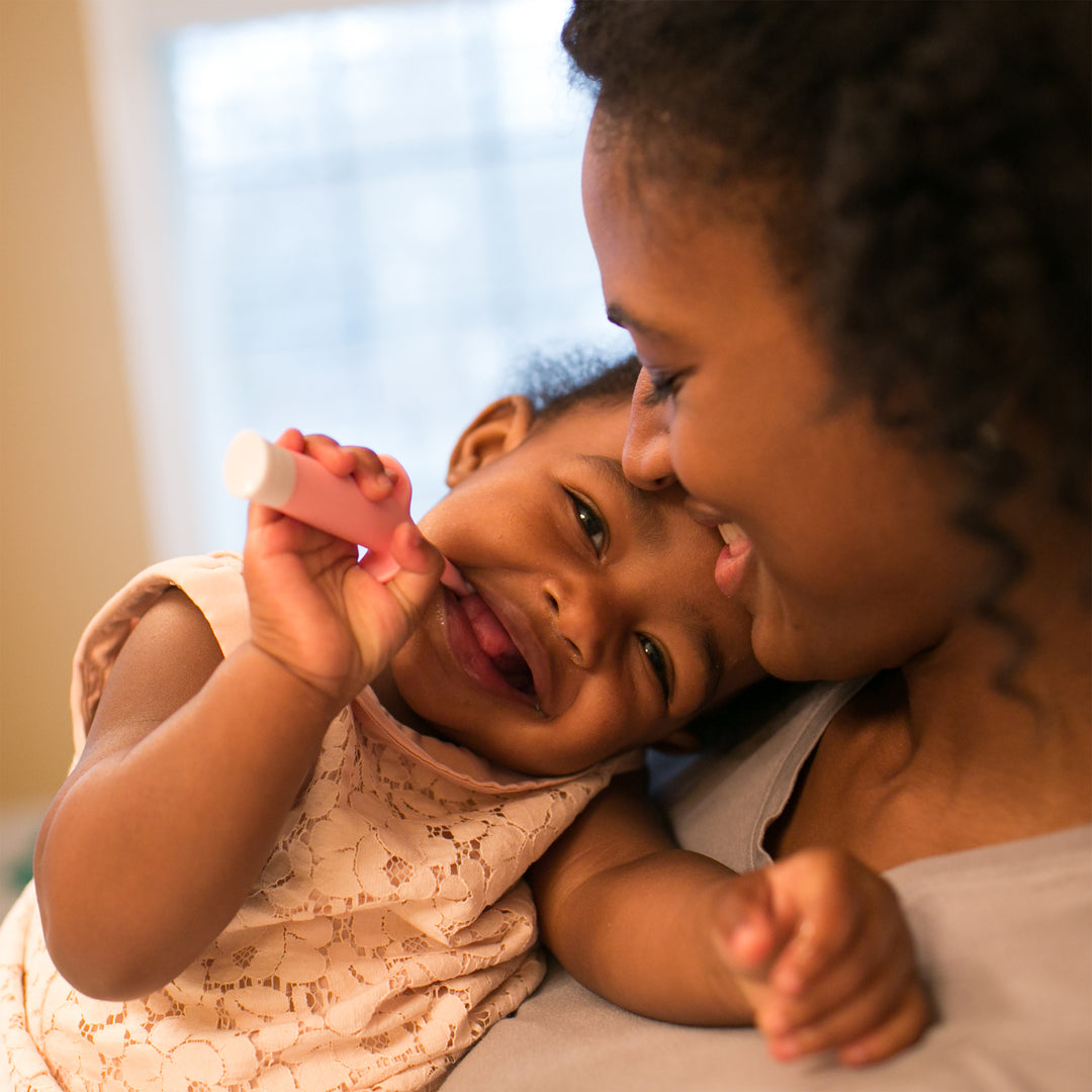 A joyful baby smiles while holding a small pink tube that looks like Dr. Brown's™ Infant-to-Toddler Toothbrush, Elephant, resting against a woman's shoulder. The woman is smiling and gently touching her nose to the baby's, creating a warm, affectionate moment in a softly lit room focused on oral hygiene.