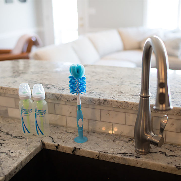 A kitchen sink with a modern faucet on a marble countertop. Beside the sink are two baby bottles featuring green accents and a Dr. Brown’s Natural Flow® Baby Bottle Brush with a stand, ideal for efficient cleaning. A blurred living room is visible in the background.