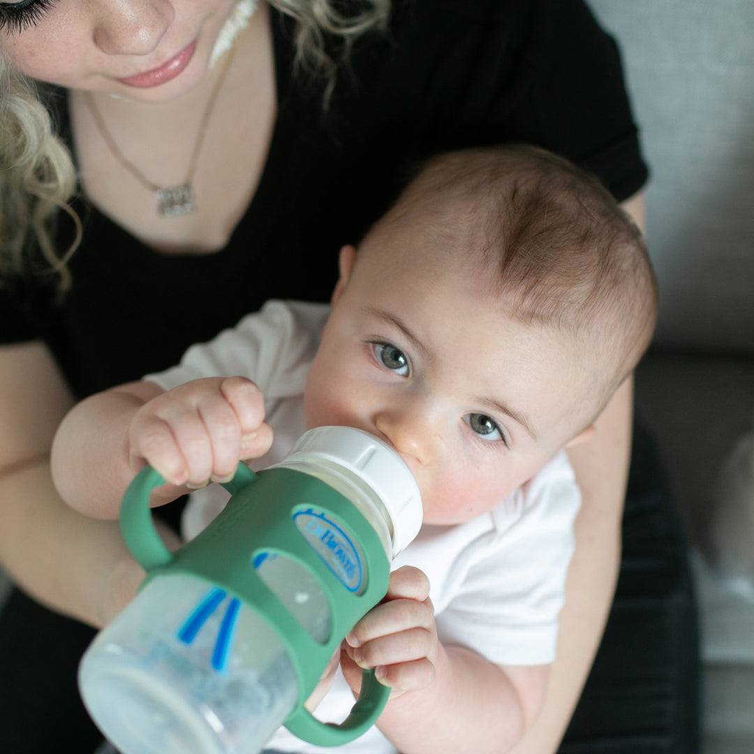 A baby with short hair and blue eyes confidently practices independent drinking skills using Dr. Brown's Milestones Wide-Neck Silicone Handles. Seated on the lap of a woman with curly blonde hair, they enjoy their time together on a gray couch, showcasing the ease granted by the no-slip easy-grip features of Dr. Brown's product.
