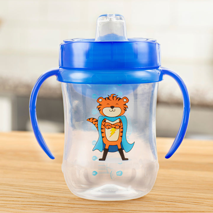 A Dr. Brown’s® Soft-Spout Toddler Cup, designed for children 9 months and older, features a blue silicone spout and handles with a playful cartoon tiger in a superhero costume on the clear cup. This 9 oz/270 ml spill-resistant cup is ideal for transitioning from bottle to cup and is pictured resting on a wooden surface against the backdrop of a softly blurred kitchen.