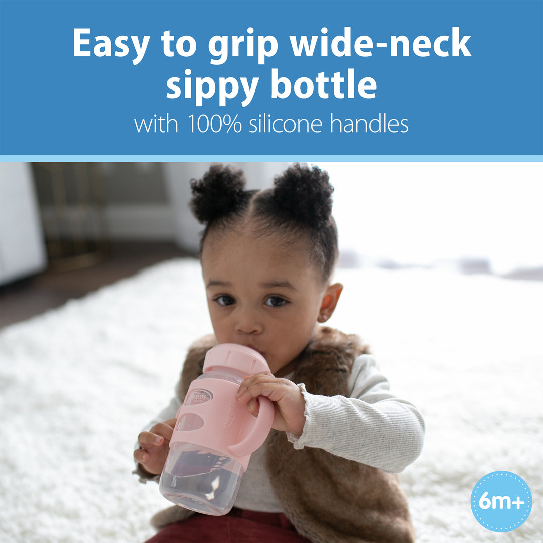 A toddler drinks from a Dr. Brown's® Milestones™ Wide-Neck Sippy Spout Bottle with Silicone Handles, 9 oz/270 mL, in pink. The text above reads, "Easy to grip for transition drinking skills," and the corner features a circle showing "6m+." The child is comfortably seated on a light-colored rug.