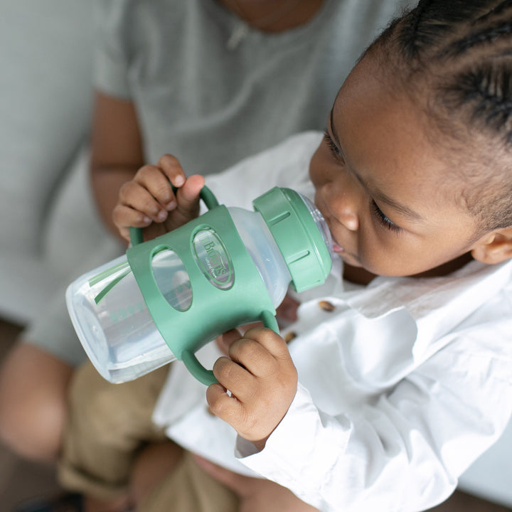 A child wearing a white shirt sips from the Dr. Brown’s® Milestones™ Wide-Neck Sippy Spout Bottle with Silicone Handles, nestled comfortably on a person's lap.