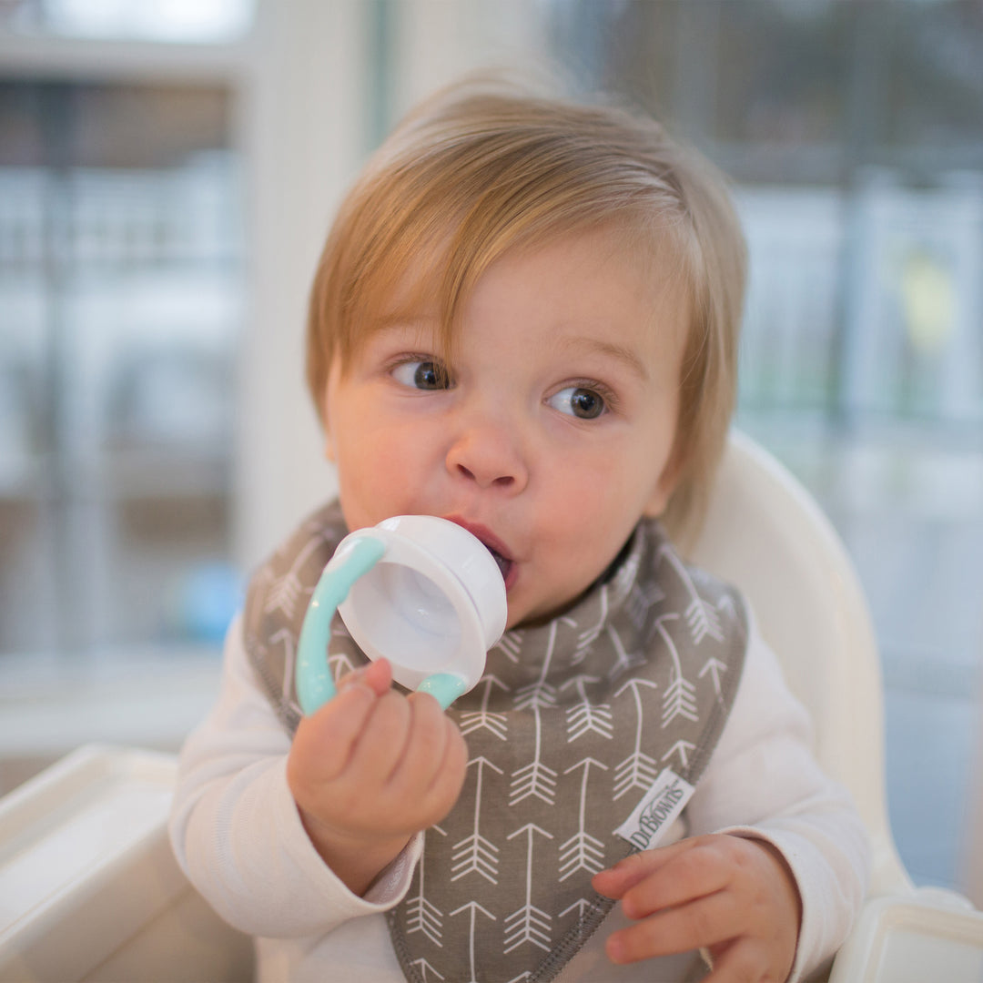 A toddler with light brown hair sits in a high chair, wearing a gray bib decorated with white arrows. The child is exploring Dr. Brown's™ Fresh Firsts™ Silicone Feeder teething toy, marking an exciting transition to solid foods, while gazing curiously to the side against a blurred indoor background.