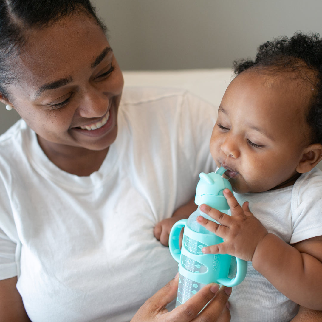 An adult wearing a white shirt with a joyful expression holds a baby, similarly dressed, on their lap. The baby is honing their independent drinking skills using the teal Dr. Brown’s™ Milestones™ Narrow Sippy Straw Bottle with Silicone Handles, 8 oz/250 mL, featuring a spill-proof lid. Both are seated indoors, creating a heartwarming moment together.