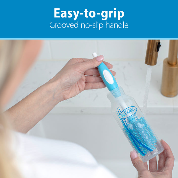 A person holds a Dr. Brown’s™ Soft Touch Bottle Brush by Dr. Brown's, featuring durable bristles and a blue grooved, easy-to-grip handle. They stand before a sink with running water, prepared to clean a baby bottle. The image emphasizes the brush's no-slip handle and its scratch-free scrubbing capabilities.