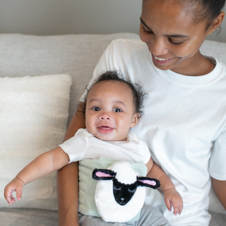 A smiling woman relaxes on a couch, holding an equally joyful baby who seems to have discovered the secret to natural gas relief with Dr. Brown’s™ Infant Gripebelt™ Colic Relief Belt. The baby wears a white outfit featuring a plush sheep decoration, and both mother and child appear content and serene in their casual white attire.