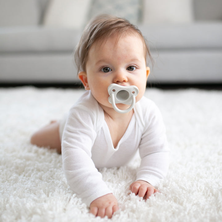 A baby with brown hair crawls across a plush white carpet, dressed in a white onesie and cheerfully sucking on Dr. Brown’s® HappyPaci™ 100% Silicone One-Piece Pacifier. A blurred gray sofa completes the cozy background scene.