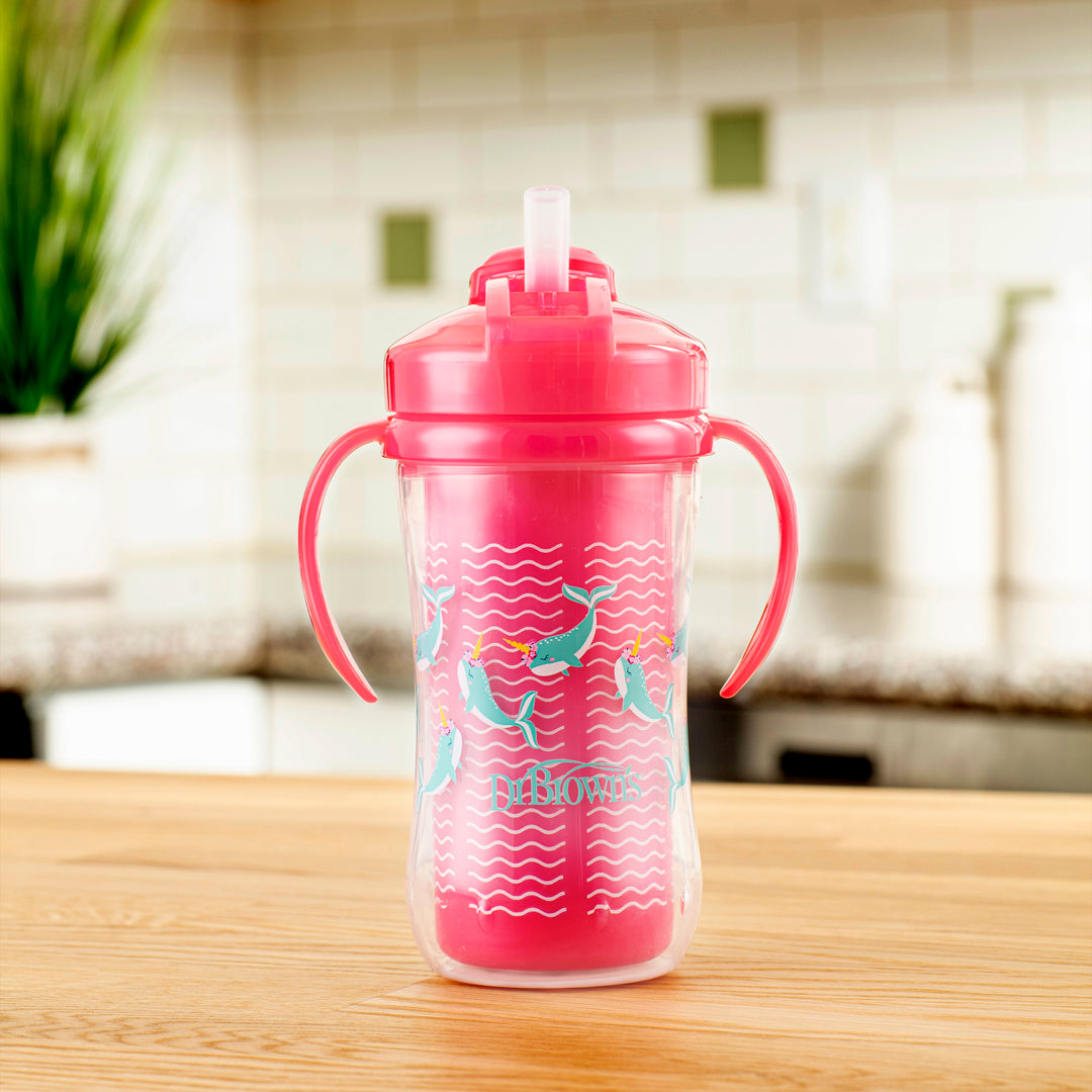 A Dr. Brown's Milestones™ Insulated Straw Cup with dual handles and a sliding lid, featuring a mermaid design, sits on a wooden kitchen counter. The pink spill-proof sippy cup is set against a blurred kitchen background with green tiles and white canisters.