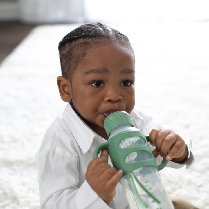 A young child with braided hair is sitting on a white carpet, wearing a white shirt, and drinking from a green Dr. Brown’s® Milestones™ Narrow Sippy Spout Bottle with Silicone Handles, 8 oz/250 mL. The child's gaze is directed forward.