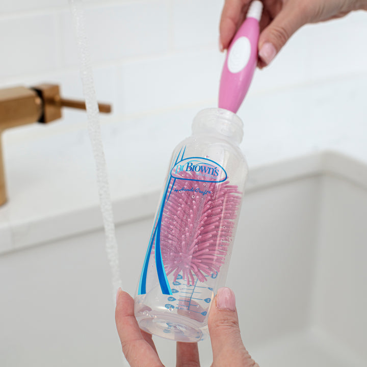 A person uses the Dr. Brown’s™ Soft Touch Bottle Brush, with its durable pink and white design, to gently scrub a baby bottle under running water in a white sink. This brush ensures scratch-free cleaning as the bottle displays the "Dr. Brown's" brand on its side.