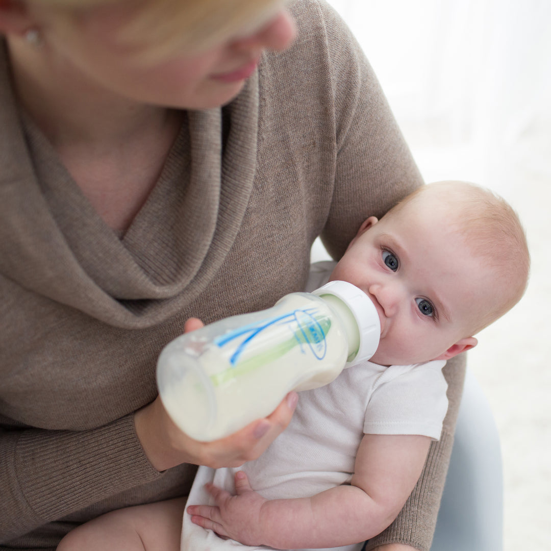 A caregiver is feeding an infant seated on their lap with a Dr. Brown's Natural Flow® Anti-Colic Options+™ Wide-Neck Baby Bottle, featuring a Level 1 Slow Flow Nipple. The baby, dressed in a white outfit, appears content while drinking from the bottle. The caregiver, wearing a beige sweater, holds the Dr. Brown's bottle gently.