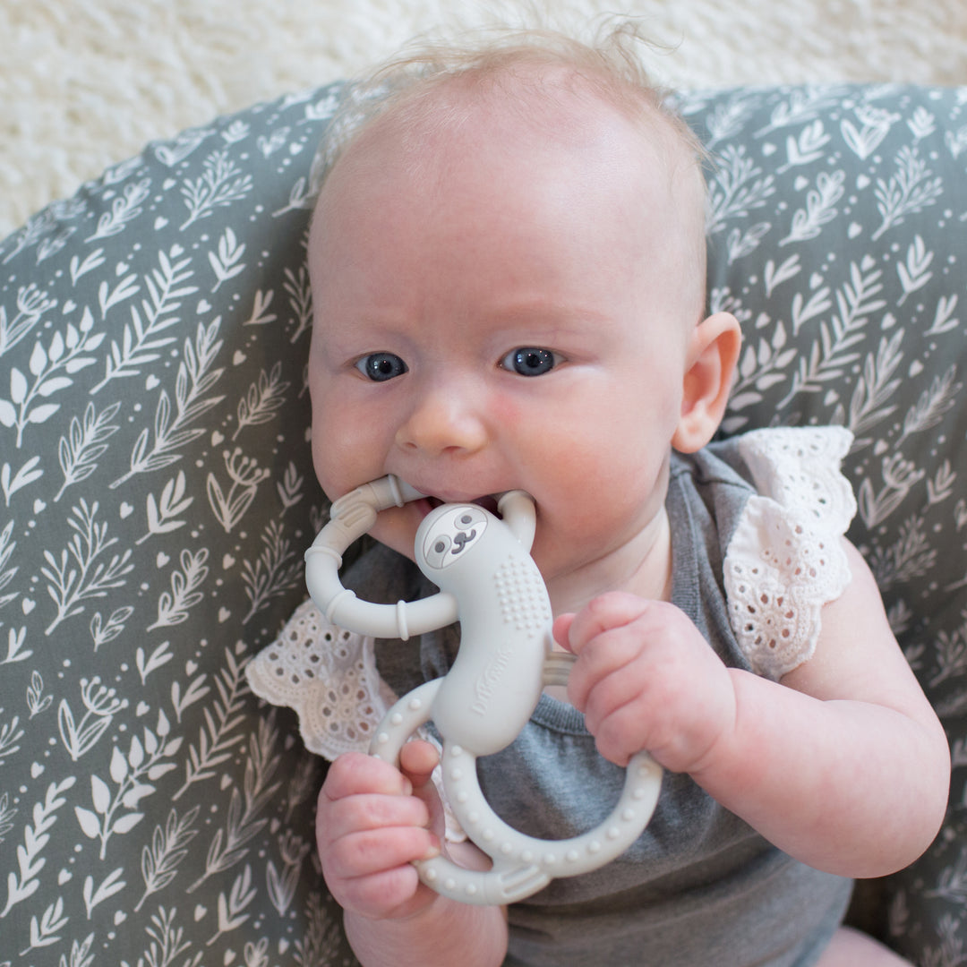 A baby with light hair is lying on a patterned cushion, dressed in a gray outfit with lace sleeves, holding and chewing on Dr. Brown's Flexees™ Sloth Teether. This silicone teether from Dr. Brown's not only soothes gums but also supports early speech development.