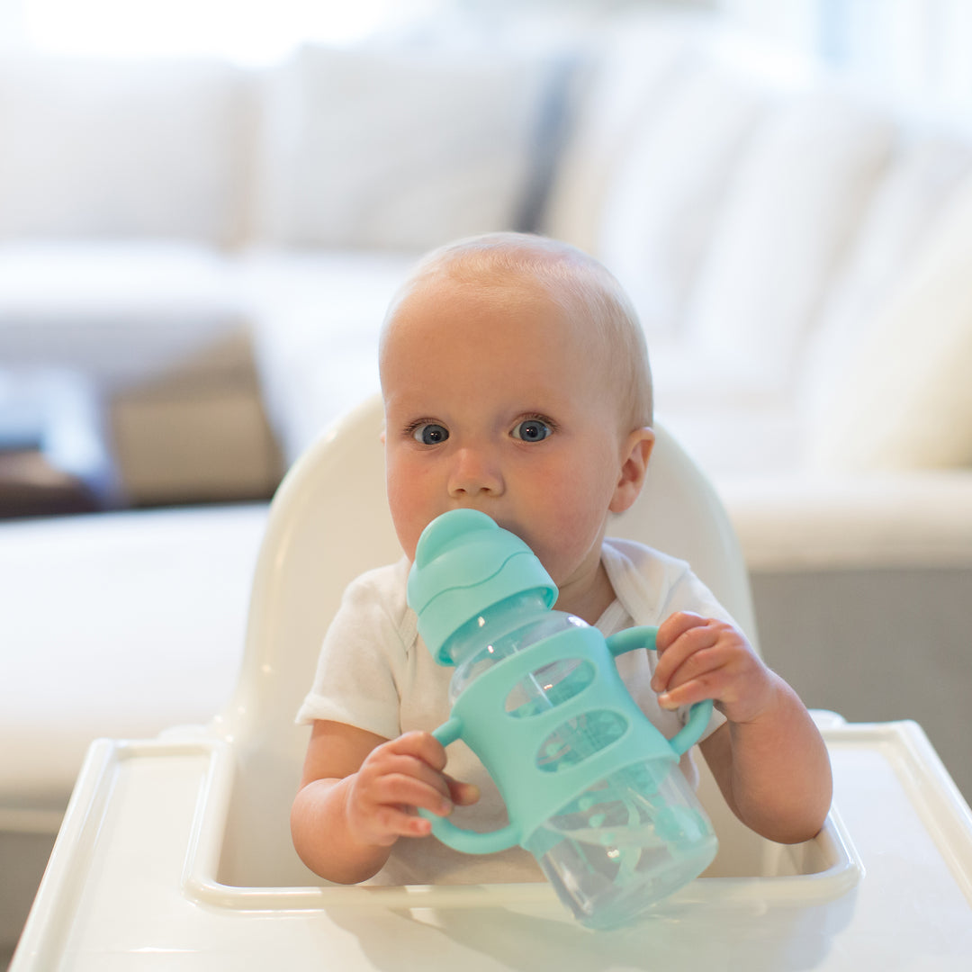 A baby sits in a high chair, independently mastering drinking skills with Dr. Brown’s™ Milestones™ Wide-Neck Sippy Straw Bottle with Silicone Handles in turquoise, holding 9 oz/270 mL. The room's soft-focus background highlights a white couch and light walls, while the silicone weighted straw ensures every sip counts.