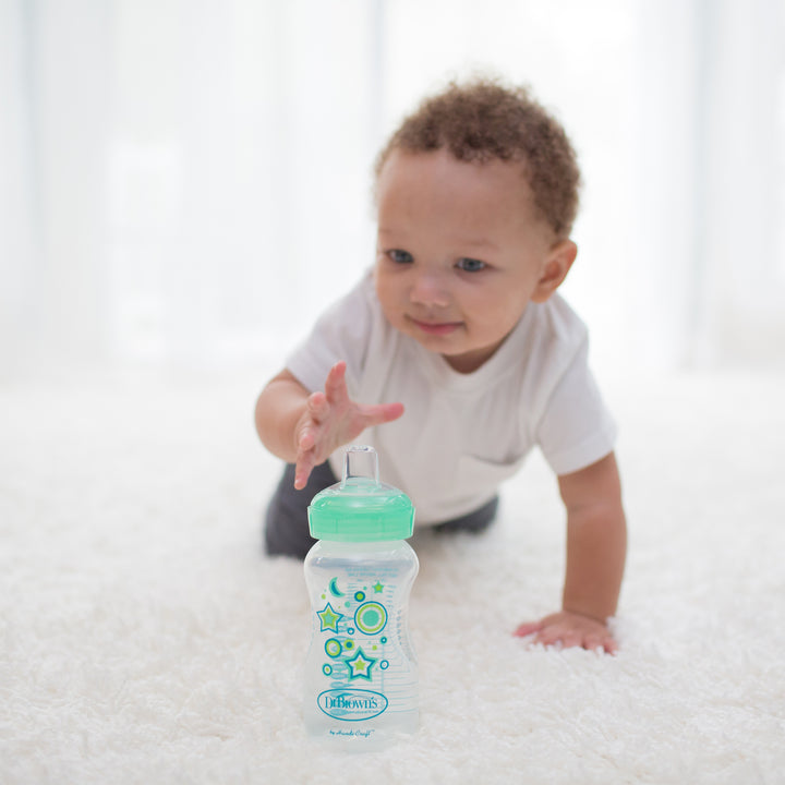 A baby in a white shirt is crawling on a soft white carpet, reaching for Dr. Brown's Natural Flow® Anti-Colic Options+™ Wide-Neck Sippy Bottle Starter Kit, 9oz/270mL, featuring a green cap and star designs. The background showcases sheer white curtains that provide soft, natural lighting.