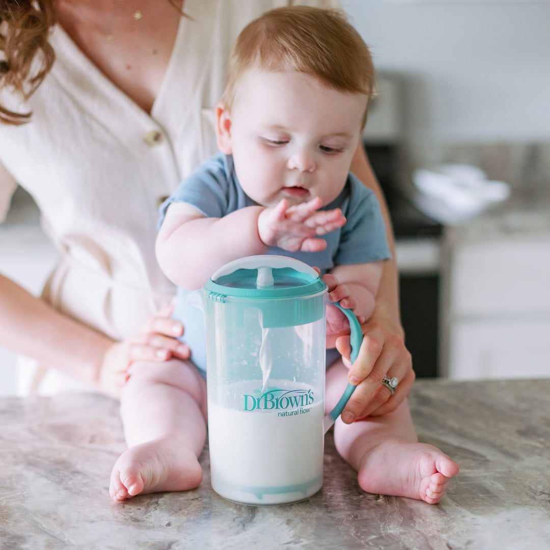 A baby perches on a kitchen counter, holding a large cup with a straw filled with milk, while an adult supports them from behind in the brightly lit scene. Nearby, Dr. Brown's Natural Flow® Formula Mixing Pitcher with its no-drip spout is stationed on the countertop.