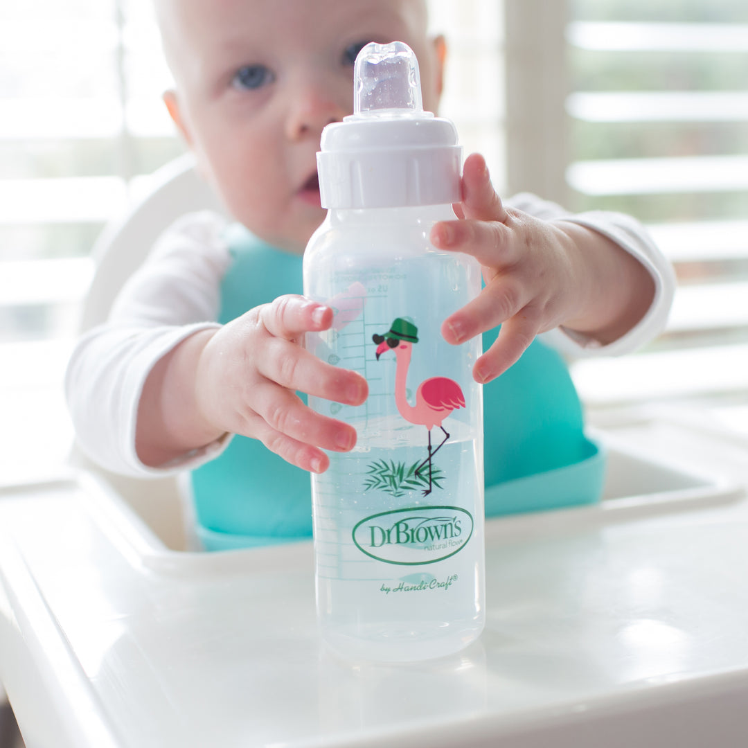 A baby in a highchair is reaching for Dr. Brown's® Milestones™ Narrow Sippy Bottle, featuring a flamingo design. Dressed in a turquoise bib, the baby is set against the gentle backdrop of windows with white blinds, preparing to transition from bottle to cup with its Silicone Sippy Spout.