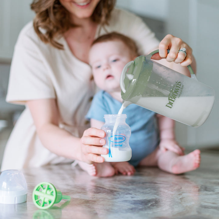 A woman is using Dr. Brown's Natural Flow® Formula Mixing Pitcher to pour milk into a baby bottle on the kitchen counter, while her baby in a blue outfit eagerly reaches for it. Nearby, various parts of Dr. Brown's baby bottles and a bottle cap are scattered across the surface.